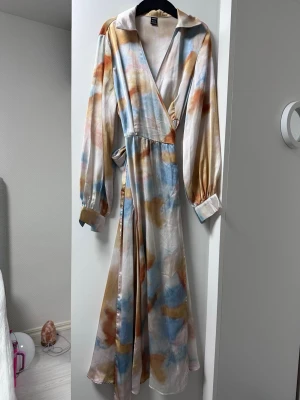 Lång omlottklänning i pastellfärger - Säljer en lång omlottklänning med pastelliga inslag av blått, orange och beige. Klänningen har långa ärmar med manschett, v-ringning och knyt i midjan. Materialet är glansigt och känns lyxigt, perfekt för dig som gillar unika plagg.