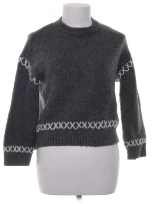 Mörkgrå stickad tröja med mönster - Mysig mörkgrå stickad tröja med vita kryssmönster runt ärmar, nederkant och bröst. Tröjan har rund halsringning, ribbade muddar och en avslappnad passform. Perfekt för kalla dagar och enkel att styla till jeans eller kjol.