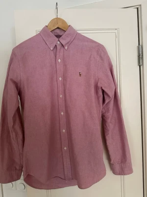 Rosa skjorta från Ralph Lauren - Klassisk rosa skjorta från Ralph Lauren med button-down krage och broderad logga på bröstet. Skjortan har långa ärmar och vita knappar framtill. Perfekt för en stilren och avslappnad look.