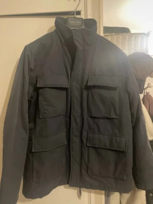 Mörkblå vindjacka från Jack's Sportswear - Snygg mörkblå vindjacka från Jack's Sportswear med fyra stora fickor framtill och hög krage. Jackan har en sportig look och är tillverkad i ett slitstarkt material som passar perfekt för blåsiga dagar. Stängs med dragkedja och tryckknappar.