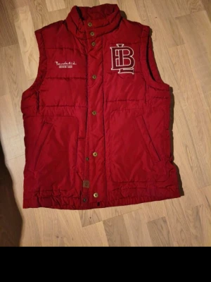 Röd dunväst från Bondelid - Riktigt snygg röd dunväst från Bondelid med hög krage, stora fickor och coola detaljer som logotypen på bröstet. Västen har knappar och dragkedja framtill och är tillverkad i ett tåligt material som håller värmen. Perfekt för lager-på-lager och streetstyle.