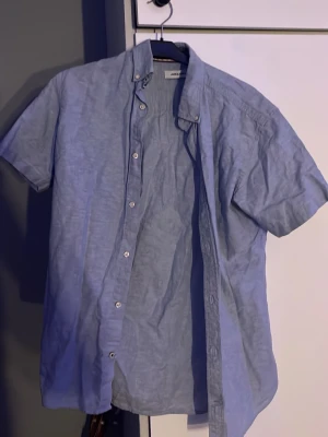Blå kortärmad skjorta från Jack&Jones - Säljer en ljusblå skjorta från Jack&Jones med korta ärmar och klassisk knappstängning. Skjortan har en relaxed passform och är tillverkad i ett lätt bomullsmaterial som känns skönt mot huden. Perfekt för dig som gillar enkel och stilren design.