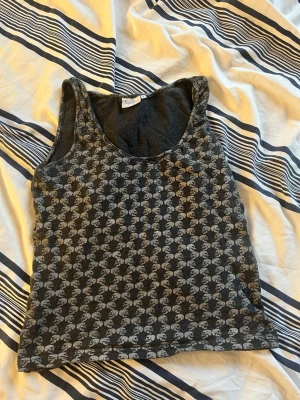 Mörkgrå mönstrad linnetopp - Snygg mörkgrå linnetopp med ljust mönster, mer exakt döskallar över hela tyget. Toppen är ärmlös och har en klassisk rund halsringning. 95% bomull & 5% lycra. Märket är björn borg.