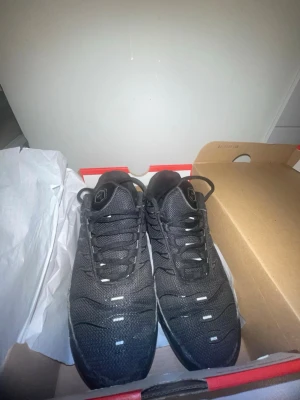 Svarta Nike Air Max Plus sneakers - Nike Air Max Plus sneakers i helsvart med mesh- och syntetmaterial, ikoniska vågiga detaljer och synlig Air-enhet i sulan. Skorna har svarta skosnören och diskreta vita accenter på ovandelen. Perfekta för dig som gillar streetwear och sportig stil.