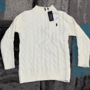 Vit Zip Knit Polo Ralph Lauren - Kabelstickad vit tröja från Polo Ralph Lauren med halv dragkedja och svart broderad logga på bröstet. Tröjan har ribbade muddar och hög krage, perfekt för en stilren och klassisk look. Materialet är mjukt och värmande. 