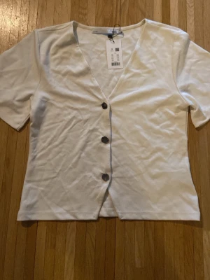 Vit kofta  - Vit v-ringad topp från 157 med korta ärmar och tre stora knappar framtill. Tillverkad i mjukt bomullsmaterial och har en enkel, clean look som passar till det mesta. Perfekt för dig som gillar minimalistisk stil.