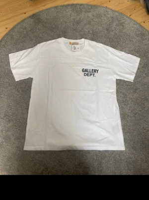 Vit Gallery Dept. t-shirt - Vit t-shirt från Gallery Dept. med svart tryck på bröstet och stor text på ryggen där det står 'GALLERY DEPT. HOLLYWOOD, CA.'. Klassisk passform och rund hals. Tillverkad i mjuk bomull som känns skön mot huden.