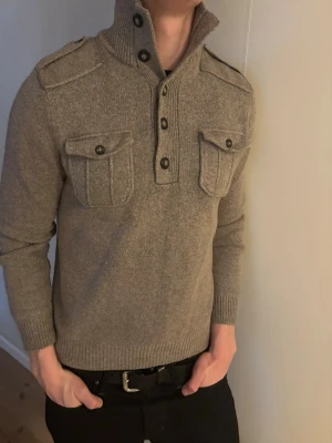 Beige brun stickad tröja med hög krage - Stickad beige tröja med hög krage och knappar framtill. Två bröstfickor med lock och knappar samt axelklaffar ger en cool militärinspirerad look. Ribbstickade muddar vid ärmslut och nederkant. Storlek m, sitter som storlek S, sitter såhär på 188cm. Mycket bra skick!