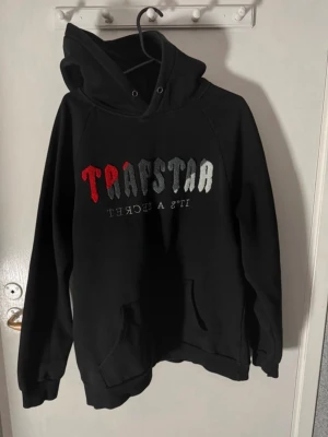 Svart Trapstar hoodie med tryck - Svart hoodie från Trapstar med stor huva och magficka. Framsidan har Trapstar-loggan i rött, grått och vitt samt texten 'It’s a secret' under. Tillverkad i mjukt bomullsmaterial som är perfekt för en avslappnad streetwear-look.