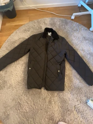 Svart vår jacka - Stilren svart vindjacka med quiltat mönster och dragkedja framtill. Jackan har två fickor med tryckknappar och klassisk krage. Perfekt för dig som vill ha en enkel men snygg jacka för våren. Märket är Bläck. Köpt för 1600 och är helt oanvänd.