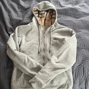 Grå hoodie från Burberry med dragkedja - Snygg grå hoodie från Burberry med dragkedja och klassiskt rutigt foder i huvan. Mjuk bomullskänsla, två fickor fram och justerbar dragsko i huvan. Perfekt för en chill och stilren look.