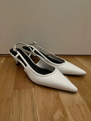 Vita slingback pumps med låg klack - Stilrena vita pumps med spetsig tå och smal rem runt hälen. Skorna har låg klack och en elegant, minimalistisk design. Perfekta för dig som gillar klassiska och trendiga skor. Materialet ser ut att vara syntet eller läderimitation.