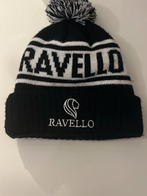 Svart och vit Ravello mössa med tofs - Säljer en svart och vit stickad mössa från Ravello med uppvikt kant och broderad logga framtill. Mössan har en stor tofs upptill och Ravello-text i vitt runt om. Perfekt för kalla dagar och snygg stil. Italienskt inspirerad mössa. 