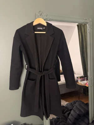Svart kappa från Pieces med skärp - Stilren svart kappa från Pieces med klassisk krage, knytskärp i midjan och två stora fickor framtill. Perfekt för kalla dagar och enkel att matcha med olika outfits. Materialet är mjukt och ger en snygg siluett. Den är lite smått nopprig🥰