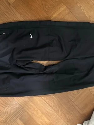 Svarta Nike mjukisbyxor med dragkedja - Säljer ett par svarta mjukisbyxor från Nike med vit logga på benet. Byxorna har dragkedja vid fickan och resår i midjan samt vid bensluten. Perfekta för chill eller träning, tillverkade i mjukt material.