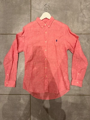 Rosa linneskjorta från Ralph Lauren - Säljer en snygg rosa linneskjorta från Ralph Lauren i slim fit-modell. Skjortan är tillverkad i linne, har långa ärmar, klassisk krage och knappar framtill. Ikoniska blå loggan broderad på bröstet. Perfekt för dig som gillar stilrena och fräscha plagg.