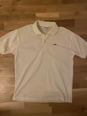 Vit pikétröja från lacoste - Klassisk vit pikétröja från Lacoste med broderad grön krokodil på bröstet. Tröjan har korta ärmar, krage och knäppning med två knappar. Tillverkad i pikétyg som ger en sportig och stilren look.