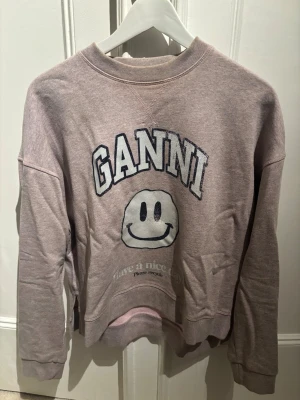  sweatshirt från Ganni - Cool ljuslila/ljusrosa sweatshirt från Ganni med tryckt smiley och texten 'GANNI' framtill. Tröjan har rund hals, ribbade muddar och avslappnad passform. Perfekt för en chill och trendig look.