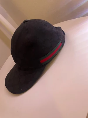Svart Gucci keps med röd-grön rand - Snygg svart keps från Gucci med diskret GG-mönster i tyget och klassisk röd-grön rand på sidan. Kepsen har böjd skärm och justerbar rem baktill. Perfekt för dig som vill ha en stilren accessoar med lyxig känsla. Priset kan diskuteras och kepsen är äkta.