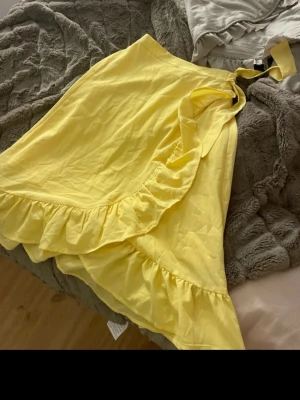 Gul omlottklänning från Lindex - Snygg gul omlottklänning från Lindex med volangdetaljer längs kanten och knyt i midjan. Klänningen har en kort längd och är perfekt för varma dagar. Tillverkad i ett mjukt och lätt material som känns skönt mot huden.