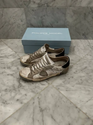 Philippe Model skor  - Snygga sneakers från Philippe Model i vitt med svarta detaljer på hälen. Skorna har en klassisk låg siluett, snörning framtill och är tillverkade i en mix av läder och mocka. Perfekta för dig som gillar en avslappnad och trendig stil. Skorna är använda och slitna framförallt under hälen därav priset. Skriv om du vill ha fler bilder eller någon fundering.👍