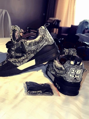 Lv skate crystal - Unika Louis Vuitton sneakers med svart och vit mönstrad textil, fransade kanter och robust svart sula. Snygga detaljer med LV-logga och grafiska element på hälen. Skorna har en chunky design och medföljer extra svarta skosnören.