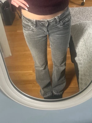 Grå bootcut jeans med låg midja - Säljer ett par gråa bootcut jeans med låg midja och slitna detaljer. Jeansen har klassiska fem fickor och dragkedja med knapp. Perfekta för en avslappnad och trendig look. Storlek 158 men uppsprättade längst ner vilket gör dem lite längre samt ger en cool effekt 💕