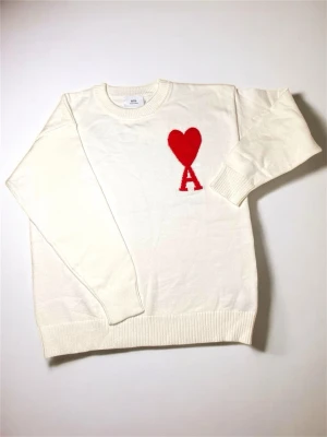 Vit stickad tröja från Ami Paris - Vit stickad tröja från Ami Paris med rött hjärta och bokstaven A broderat på bröstet. Tröjan har rund halsringning, ribbade muddar och nederkant. Perfekt för dig som gillar stilrena och trendiga plagg.