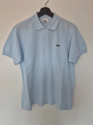 Ljusblå pikétröja från Lacoste - Klassisk ljusblå pikétröja från Lacoste med korta ärmar och den ikoniska gröna krokodilloggan på bröstet. Tr download har krage och knäppning med två knappar framtill. Perfekt basic till garderoben sopassad för en clean och stilren look.