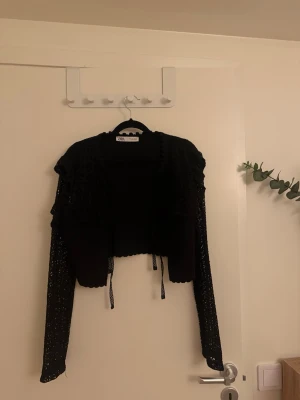 Svart virkad kofta från Zara - Snygg svart virkad kofta från Zara med volangdetaljer och knytband framtill. Koftan är kortare i modellen och har långa ärmar med ett genombrutet mönster som ger en trendig look. Perfekt att slänga över en topp för en cool outfit. (Liten i storleken)