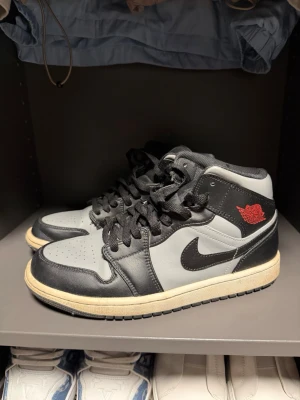 Air Jordan 1 Mid svart/grå - Snygga Air Jordan 1 Mid sneakers i svart och grått med klassisk röd Air Jordan-logga på sidan. Skorna har svart swoosh, svarta skosnören och en beige sula. Tillverkade i läder med textildetaljer och ikonisk siluett.