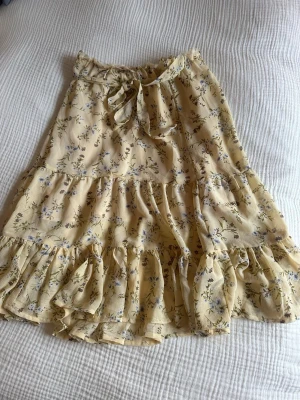 Gul blommig volangkjol med knytband - Söt gul kjol med blommigt mönster och volangdetaljer. Kjolen har knytband i midjan och är tillverkad i ett lätt, luftigt material. Knälängd. 