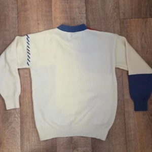 Flerfärgad stickad tröja med patchwork - Unik stickad tröja i vitt, blått och orange med patchwork-design. Tröjan har blå ribbstickad krage och ärmslut, samt dekorativa sömmar och kabelstickade partier. Perfekt för dig som vill sticka ut med en kreativ och färgglad stil. 