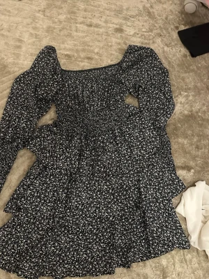 Blommig svart kortklänning - Supersöt svart kortklänning med vita små blommor och långa puffärmar. Klänningen har smockad överdel och volangdetaljer i kjolen, vilket ger en romantisk vibe. 