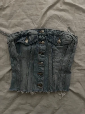 Jeans korsettopp med knappar - Snygg korsettopp i denim med knappar framtill och rå kant nertill. Toppen har dekorativa fickor med knappdetaljer och tunna axelband. Perfekt för dig som vill ha en edgy och trendig look.
