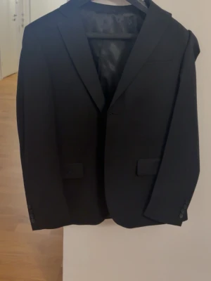 Svart kavaj Dressmann Slim Fit - Stilren svart kavaj från Dressmann i modellen Slim Fit. Kavajen har klassisk krage, två framfickor med lock och knappar vid ärmsluten. Fodret är svart med röda detaljer. Perfekt för en dressad look. Köpt maj 2024, används 2 gånger, inga synliga fläckar. Både kavaj och kostymbyxor ingår. 