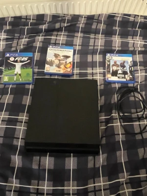 PlayStation 4 med spel och kablar - Säljer en svart PlayStation 4 från Sony med stilren, rektangulär design. Konsolen är tillverkad i plast och kommer med tre PS4-spel: FC 25, Overwatch och UFC samt tillhörande strömkabel. Perfekt för gaming och multiplayer.
