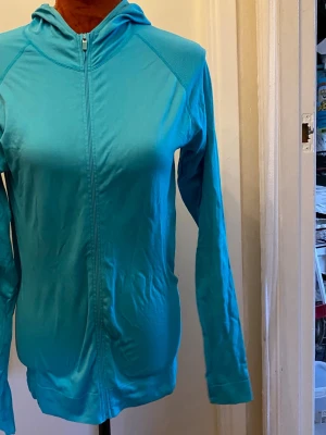 Turkos träningshoodie med dragkedja M - Träningströja med hel dragkedja framtill och huva. Långa ärmar med raglanärmskärning. Tröjan har en figurnära passform. Längd: 67 cm Bystvidd: 42 cm  Inga bilder när den sitter på mig! Katter finns i hemmet!