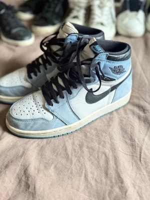 Nike Air Jordan 1 High University Blue - Snygga Nike Air Jordan 1 High i färgen University Blue med svart swoosh och svarta skosnören. Ovandelen är i vitt och ljusblått läder och mocka, med klassisk Air Jordan-logga på sidan. Perfekta sneakers för dig som gillar streetwear och vill sticka ut. OBS lådan till skorna är sönder. 