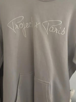 Grå hoodie Project X Paris - Snygg grå hoodie från Project X Paris med broderad logga framtill. Hoodien har en stor känguruficka och en klassisk huva. Tillverkad i mjukt material som känns skönt mot huden. Perfekt för en avslappnad och trendig look.