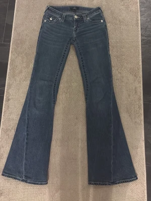 True Religion utsvängda jeans blå - Säljer ett par blå utsvängda jeans från True Religion med klassiska kontrastsömmar och dekorativa fickor bak med lock och knappar. Jeansen har låg midja.