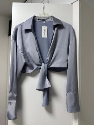Blå/Silvrig satinblus - oanvänd - Trendig silvrig satinblus med lång ärm och knytning framtill. Blusen har klassisk krage och breda manschetter med knappdetalj. Den korta modellen ger en cool och modern look, perfekt att styla med höga jeans eller kjol.
