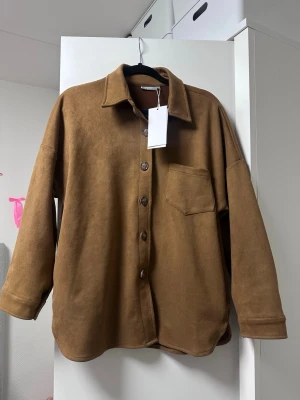Brun overshirt med ficka (läder immitation) - Snygg brun overshirt med klassisk krage och knäppning framtill. Modellen har en bröstficka och är tillverkad i ett mjukt, mockaliknande material. Perfekt för lager-på-lager och ger en chill vibe till din outfit.