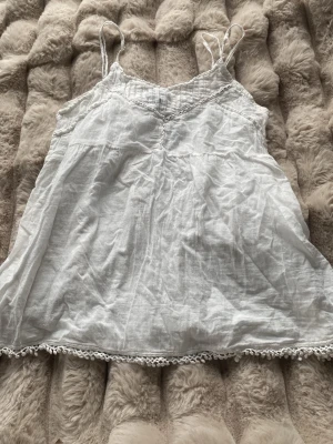 Vit linnetopp med spetsdetaljer - Söt vit linnetopp med tunna axelband och spetsdetaljer längs halsringning och nederkant. Toppen är luftig och har en avslappnad passform, perfekt för varma dagar. Materialet känns lätt och toppen har fina virkade detaljer längst ner.