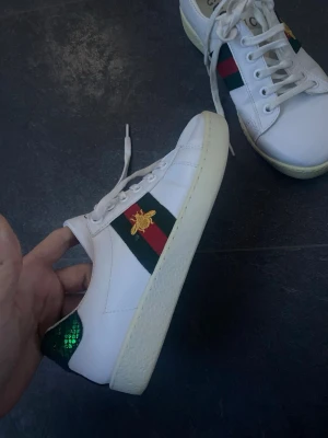 Sneakers skor 👟 från G - Vita sneakers i G-inspirerad stil med klassiska grön/röda ränder på sidan och en snygg guldbroderad detalj 🐝✨ Supertrendiga och passar till allt – jeans, kostymbyxor eller klänning. Sköna och stilrena med grön glittrig detalj baktill som ger det lilla extra 💚  Fint skick (använda men väl omhändertagna). Perfekta för dig som vill ha en lyxig look till ett bra pris!