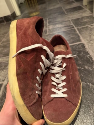 Vinröda sneakers från Golden Goose - Säljer ett par vinröda sneakers från Golden Goose med vit sula och vita skosnören. Skorna är i mocka och har den klassiska stjärndetaljen på sidan. Perfekta för dig som gillar en avslappnad och trendig stil.