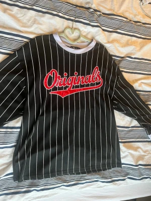 Svart randig långärmad tröja Originals - Svart långärmad tröja med vita vertikala ränder och stor röd broderad text 'Originals' på bröstet. Tröjan har vita kanter vid hals och ärmslut för en lite sportig look. Har två minimala skador  (se sista bilderna), ena är lagad men skulle säga knappt märktbart eller svårt och fixa. Material okänt för avklippt lapp.