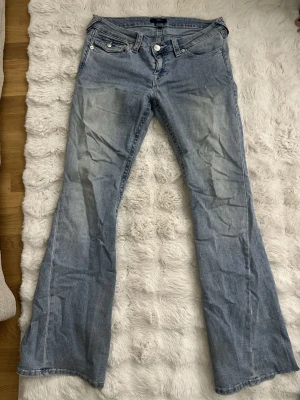 Ljusblå flared jeans från True Religion - Säljer ett par ljusblå flared jeans från True Religion med kontrastsömmar och vita knappar. Jeansen har låg midja.