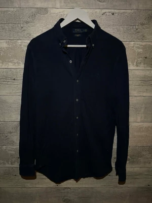 Mörkblå skjorta från Polo Ralph Lauren - Klassisk mörkblå skjorta från Polo Ralph Lauren med button-down krage och knappar framtill. Skjortan har lång ärm och en stilren, tidlös design som passar till många olika outfits.