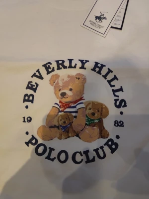 Vit t-shirt Beverly Hills Polo Club - Vit t-shirt från Beverly Hills Polo Club med ett stort tryck av tre nallebjörnar på bröstet. T-shirten har rund hals och korta ärmar. Trycket är färgglatt med blå, röd och grön detalj. Perfekt för dig som gillar lekfulla prints och klassisk stil.